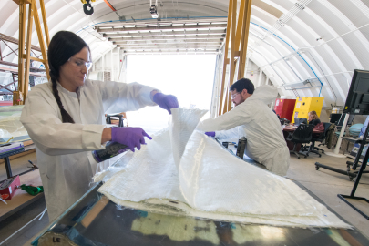 Two people in safety attire adjust fabric layers in a manufacturing facility